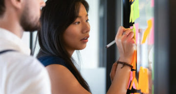 Focused asian business woman mentor coach leader writing idea or task on post it sticky notes on glass wall, serious team people developing work plan in creative corporate office at stand up meeting